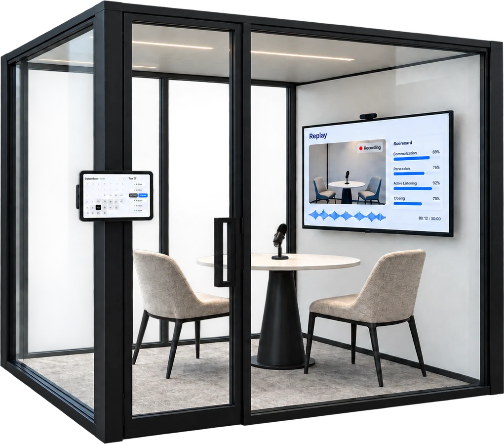 A glass role-play booth with a table, two chairs, a microphone, a TV showing a Replay recording and scorecard, and a booking tablet by the door